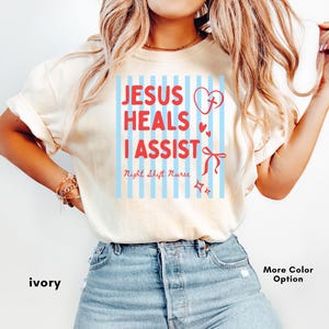 Puede incluir: Una camiseta de manga corta color marfil con un gráfico de rayas verticales azul claro y texto en rojo que dice "JESUS HEALS I ASSIST" y "Night Shift Nurse". El diseño incluye un corazón rojo con una cruz, corazones pequeños, un lazo y estrellas.