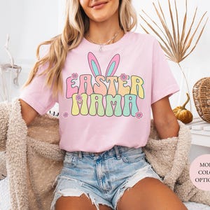 Puede incluir: Camiseta rosa claro con la frase "EASTER MAMA" en colores pastel y orejas de conejo. La modelo lleva pantalones cortos vaqueros. Un cárdigan beige está sobre los hombros.
