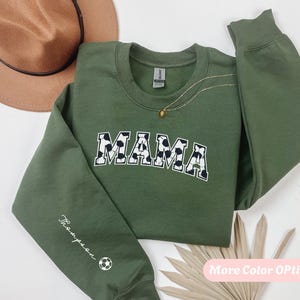 Puede incluir: Una sudadera verde oliva con la palabra "MAMA" en un diseño de estampado de vaca en blanco y negro. La manga tiene el nombre "Thompson" y un gráfico de balón de fútbol. Se ve un collar dorado.