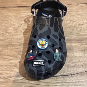 Puede incluir: Zapato Crocs de camuflaje negro y gris con una correa negra. El zapato presenta varios dijes decorativos, incluido un logotipo del Manchester City y un dije con el texto "oasis".
