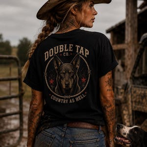 May include: A black t-shirt with the text "DOUBLE TAP CO." and a dog graphic. The shirt is worn with blue jeans and a cowboy hat. A black and white dog is in the foreground.