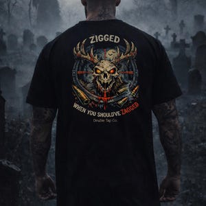 Puede incluir: Camiseta negra con un diseño de calavera con cuernos, una mira y balas. El texto "ZIGGED WHEN YOU SHOULD'VE ZAGGED" está arriba y abajo del gráfico. La camiseta la lleva una persona en un cementerio.