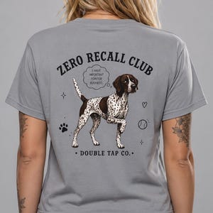 Può includere: Una t-shirt grigia a maniche corte con la grafica di un cane maculato marrone e bianco. Il testo recita ZERO RECALL CLUB, DOUBLE TAP CO., e un fumetto dice I HAVE IMPORTANT POINTER BUSINESS. Include piccole icone di una zampa, cuore e palla.