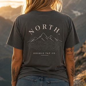 May include: A charcoal gray short-sleeve t-shirt featuring a white graphic on the back. The design includes the word NORTH in an arc above a line-art mountain range and topographic lines, with the text DOUBLE TAP CO. printed below the graphic.