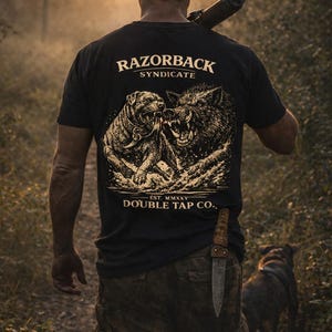 May include: Black t-shirt with a graphic of a dog and a boar fighting, with the text "RAZORBACK SYNDICATE" and "DOUBLE TAP CO." A person is walking in a forest, carrying a rifle and a knife.