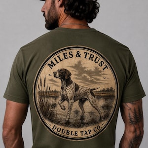 Pode incluir: Camiseta verde-oliva com um gráfico circular apresentando um cão de caça em um pântano. O gráfico inclui o texto "MILES & TRUST" e "DOUBLE TAP CO.". A pessoa está usando um boné marrom.