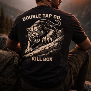 Puede incluir: Camiseta negra con un diseño blanco de un felino rugiendo sobre una roca, con el texto "DOUBLE TAP CO." y "KILL BOX". La persona lleva la camiseta al aire libre, con un paisaje montañoso.
