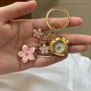 Peut inclure: Porte-clés doré avec une montre en forme de cœur et des breloques florales. Le cadran de la montre a des chiffres et des aiguilles noirs. Les breloques comprennent trois petites fleurs rose clair et une plus grande fleur rose avec un centre doré.