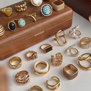 Puede incluir: Una colección de anillos dorados con diversos diseños, incluyendo formas de corazón, adornos de perlas y estilos geométricos. Algunos anillos presentan piedras preciosas y detalles texturizados. Los anillos se exhiben sobre una caja de madera y una superficie blanca.