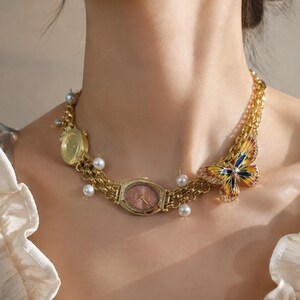 Peut inclure: Un collier à chaîne dorée composé de deux cadrans de montre ovales intégrés, dont un avec un fond rose, agrémenté de perles sphériques blanches et d'un papillon en émail multicolore aux ailes bleues, jaunes et orange avec des strass.