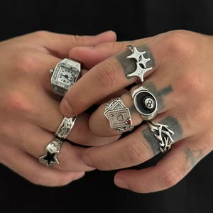 Op de afbeelding: Een verzameling zilveren ringen met verschillende ontwerpen, waaronder een ring in de vorm van een horloge, een ster, een speelkaart en een ring met een achtbal. De ringen worden op meerdere vingers gedragen.