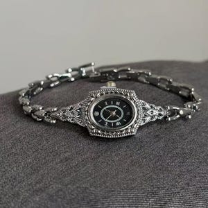 Op de afbeelding: Een zilverkleurig polshorloge met een ovale zwarte wijzerplaat en Romeinse cijfers. Het horloge heeft een sierlijke lunette en een schakelarmband. Een kleine diamant is op 6 uur geplaatst.