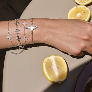 May include: A collection of silver bracelets with cross and gemstone designs. The bracelets are displayed on a person's wrist, alongside sliced lemons on a neutral-toned surface. The jewelry features delicate chains and intricate cross details.