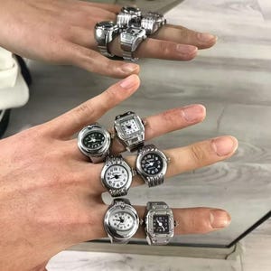 以下が含まれることがあります： 丸、楕円、正方形など、さまざまな文字盤デザインのシルバーカラーのリングウォッチのコレクション。時計は黒と白の文字盤で、一部にはローマ数字が使用されています。指にリングが表示されています。