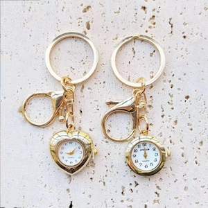 May include: Two gold-colored keychains with small quartz watches. One watch is heart-shaped, the other is round. Both have white faces with black numerals and hands. The keychains have a clasp and a ring.
