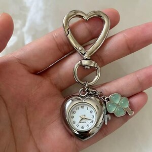 May include: Silver heart-shaped pocket watch with a white clock face and black numerals. The watch has a matching heart-shaped clasp and a charm of a four-leaf clover. The watch is attached to a silver heart-shaped clip.