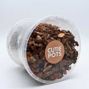 May include: A clear plastic container filled with brown bark chips. A circular sticker with the words "CUTIE POTS" in white letters is affixed to the container. The container is tilted, revealing the contents.