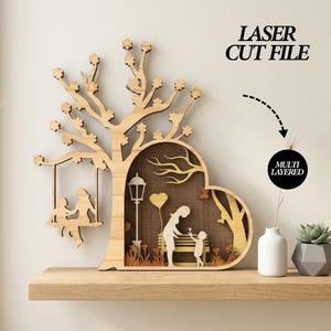 Könnte beinhalten: Mehrschichtige Laserdatei aus Holz, die einen Baum mit einer Schaukel und einem herzförmigen Ausschnitt darstellt. Der Ausschnitt zeigt eine Mutter und ein Kind, eine Parkbank und eine Straßenlaterne. Der Text "LASER CUT FILE" ist sichtbar.