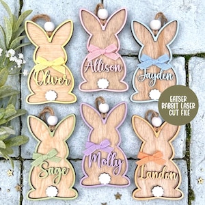 Könnte beinhalten: Osterhasen-Ornamente aus Holz mit personalisierten Namen und bunten Schleifen. Jeder Hase hat einen weißen Pompon-Schwanz und eine Schnurschlaufe zum Aufhängen. Namen: Oliver, Allison, Jayden, Sage, Molly und Landon. Der Text "EATSER RABBIT LASER CUT FILE" ist ebenfalls sichtbar.