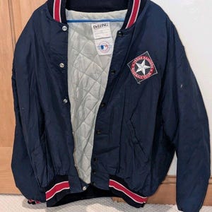 May include: Navy blue DeLong baseball jacket with a quilted lining. The jacket has red and white striped detailing on the collar, cuffs, and hem. A Texas Rangers Baseball Club patch is on the chest.