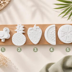 Könnte beinhalten: Fünf weiße, mattierte Formen, die auf einem rechteckigen Holzbrett angeordnet sind. Die Formen umfassen einen Flamingo, eine Ananas, eine Erdbeere, eine Wassermelonenscheibe und eine Zitrusfruchtscheibe. Jede Form ist mit eins bis fünf nummeriert.