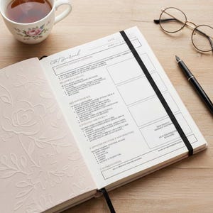 Pode incluir: Um diário aberto com uma faixa elástica preta, uma xícara de chá, óculos e uma caneta em uma superfície de madeira. A página esquerda do diário tem um design floral em relevo, enquanto a página direita tem texto e caixas. O diário é rotulado como "CBT Journal".