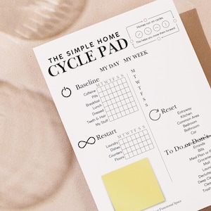 May include: A white "CYCLE PAD" notepad with the text "THE SIMPLE HOME" and "MY DAY MY WEEK" printed on it. The notepad has sections for "Baseline", "Restart", "Reset", and "To Do or Don'ts". A yellow sticky note is attached. Two glasses of water are in the background.