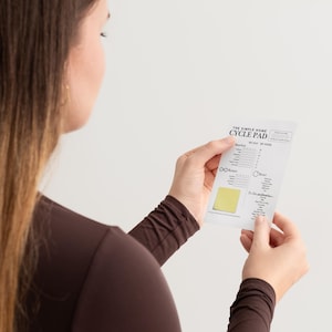 May include: A white cycle pad with the text "THE SIMPLE HOME CYCLE PAD" is held by a person. The pad features a yellow sticky note and a calendar layout with symbols and text. The person wears a brown long-sleeved top.
