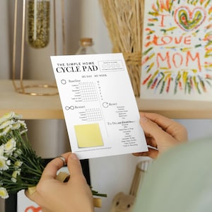 May include: A person holds a white "THE SIMPLE HOME CYCLE PAD" with a yellow sticky note. The pad has sections for "MY DAY" and "MY WEEK" with checkboxes. A colorful "I LOVE MOM" drawing is in the background.
