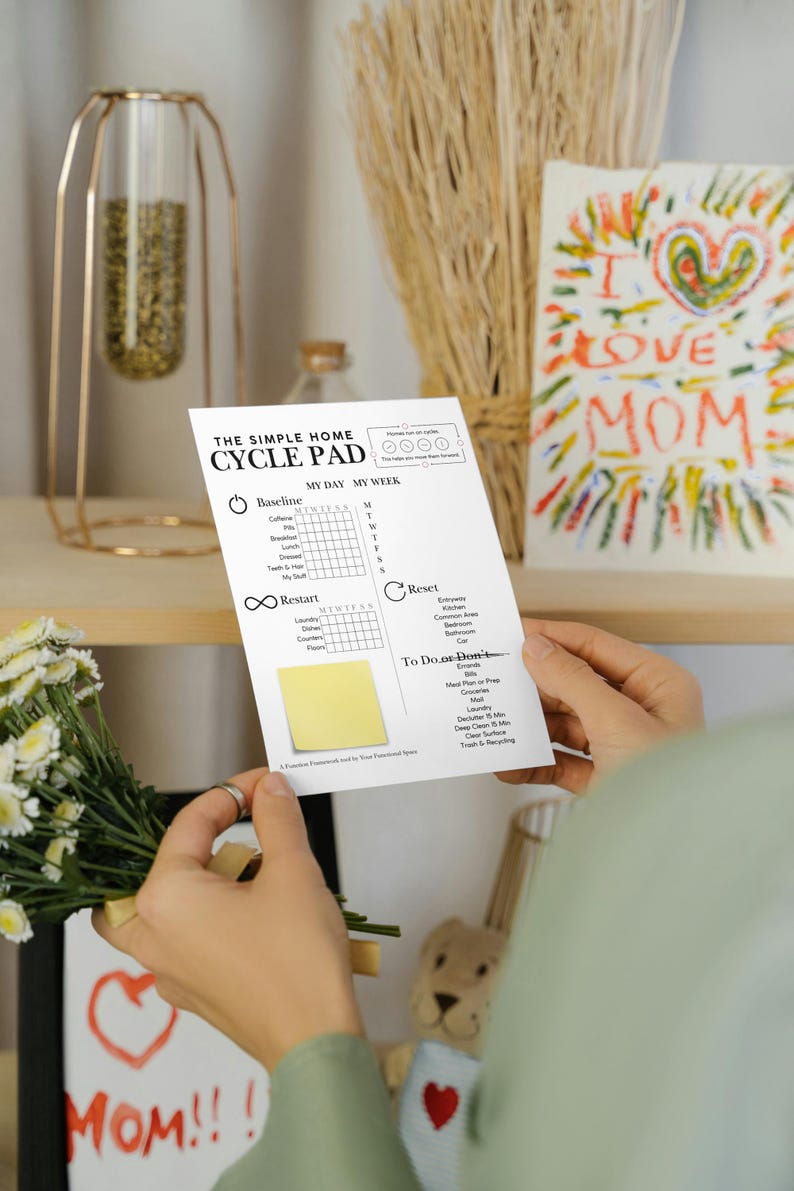May include: A person holds a white "THE SIMPLE HOME CYCLE PAD" with a yellow sticky note. The pad has sections for "MY DAY" and "MY WEEK" with checkboxes. A colorful "I LOVE MOM" drawing is in the background.
