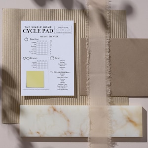 May include: A flat lay composition featuring a white and beige cycle pad with the text "THE SIMPLE HOME CYCLE PAD". The pad is on a corrugated cardboard background, with a beige ribbon, a brown square, and a marble-like rectangular block.