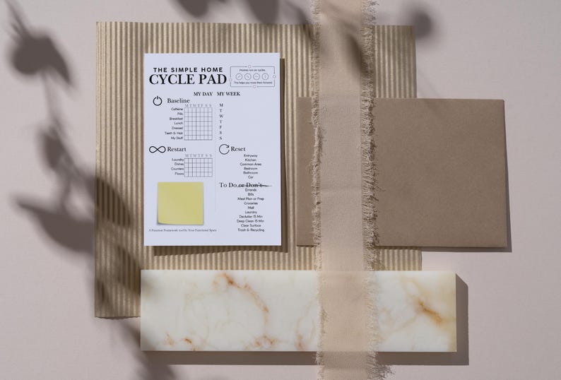 May include: A flat lay composition featuring a white and beige cycle pad with the text "THE SIMPLE HOME CYCLE PAD". The pad is on a corrugated cardboard background, with a beige ribbon, a brown square, and a marble-like rectangular block.