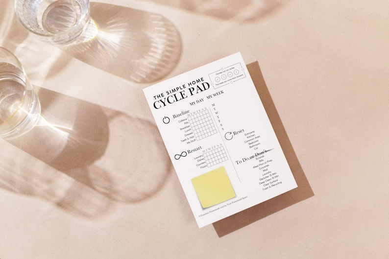 May include: A white "CYCLE PAD" notepad with the text "THE SIMPLE HOME" and "MY DAY MY WEEK" printed on it. The notepad has sections for "Baseline", "Restart", "Reset", and "To Do or Don'ts". A yellow sticky note is attached. Two glasses of water are in the background.