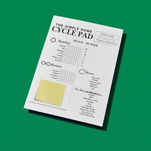 May include: A white paper pad with the text "THE SIMPLE HOME CYCLE PAD" and a yellow sticky note. The pad includes sections for daily and weekly tracking of habits and tasks, with a green background.