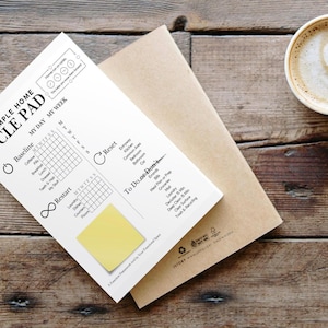 May include: A white "CYCLE PAD" notepad with a yellow sticky note, on a brown envelope. The notepad features a chart for daily and weekly tracking. A coffee cup with latte art is placed on a wooden surface.