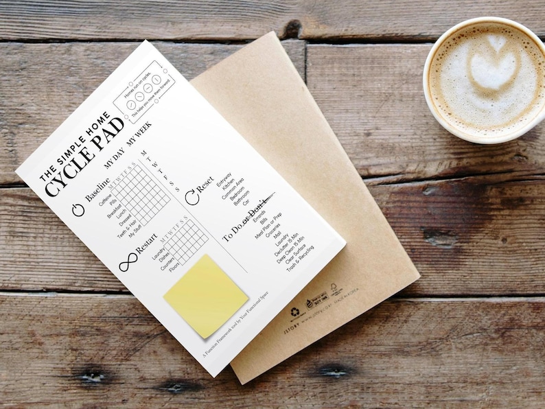 May include: A white "CYCLE PAD" notepad with a yellow sticky note, on a brown envelope. The notepad features a chart for daily and weekly tracking. A coffee cup with latte art is placed on a wooden surface.