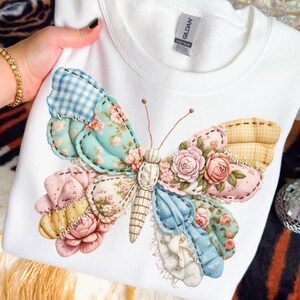 Può includere: Felpa bianca con un grande e colorato motivo a farfalla. Le ali della farfalla sono patchwork con motivi floreali e a quadretti, e il corpo ha un aspetto cucito. Il design incorpora colori rosa, blu, giallo e bianco.