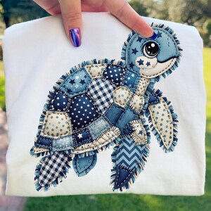 May include: A white sweatshirt featuring a patchwork turtle design. The turtle is made of various blue fabrics with star and polka dot patterns. The turtle's head and flippers are detailed with stitched edges.