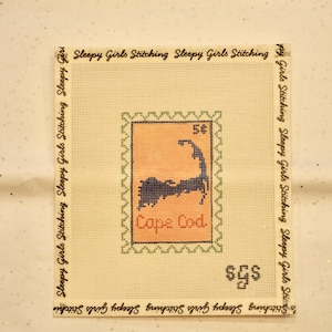 May include: A needlepoint canvas featuring a Cape Cod design within a postage stamp-like frame. The design is stitched in blue and orange hues, with the text "Cape Cod" below. The canvas is surrounded by the text "Sleepy Girls Stitching".