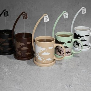 May include: Five ceramic mugs with matching saucers, each featuring a different color and whale design. The mugs are black, brown, tan, mint green, and white. Each mug has a tea bag holder attached.