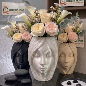 May include: Three decorative vases shaped like faces, each holding a floral arrangement. The vases are black, pearlescent white, and beige. The flowers are roses, calla lilies, and other greenery. The vases are on a dark surface.