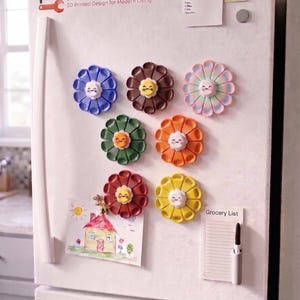 May include: Six colorful flower-shaped magnets with smiling faces attached to a white refrigerator. The magnets are blue, brown, pastel rainbow, green, orange, red, and yellow. A logo reads HACKING HOME 3D Printed Design for Modern Living.