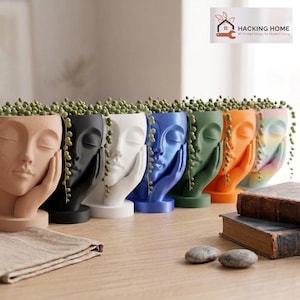 May include: A row of seven face-shaped planters in matte beige, black, white, blue, green, orange, and a multi-colored finish. Each planter features a sculpted face resting on a hand and contains a trailing string of pearls plant.