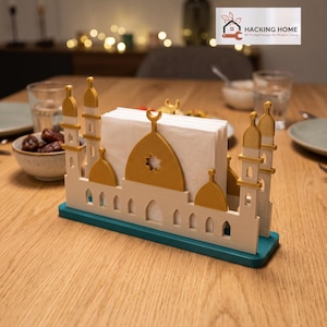 May include: A 3D printed napkin holder shaped like a mosque with a teal base, cream-colored walls, and gold-colored domes and minarets. The structure features a central arch with a star cutout and holds a stack of white paper napkins.