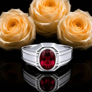 Könnte beinhalten: Ein silberfarbener Ring mit einem markanten, oval geschliffenen, tiefroten Edelstein, gefasst in eine polierte Metallschiene mit gestuften Seitendetails. Der Ring ist vor drei blass pfirsichfarbenen Rosen auf schwarzem Grund platziert.