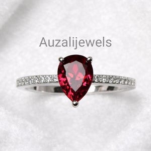 Op de afbeelding: Een zilveren ring met een peervormige, dieprode edelsteen als middelpunt. De band is versierd met kleine, heldere edelstenen. De tekst "Auzalijewels" wordt grijs over de bovenkant van de afbeelding weergegeven. Deze ring is een fijn sieraad.