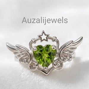 May include: A silver ring featuring a heart-shaped, light green gemstone. The ring has a star and wing design, with the text "Auzalijewels" in the background. The ring is set against a soft, white background.