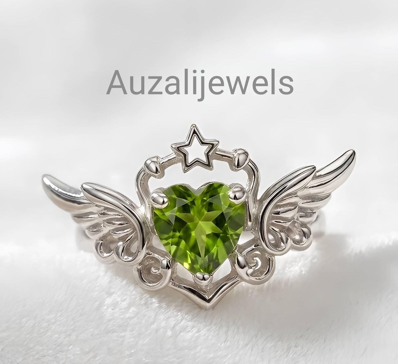 May include: A silver ring featuring a heart-shaped, light green gemstone. The ring has a star and wing design, with the text "Auzalijewels" in the background. The ring is set against a soft, white background.