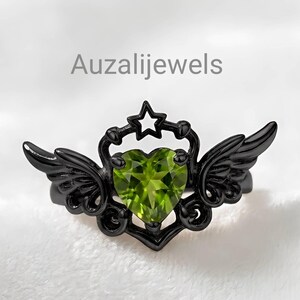 May include: Black ring with a heart-shaped, light green gemstone. The ring features a star and wing design. The text "Auzalijewels" is visible on the image.