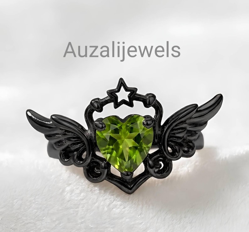 May include: Black ring with a heart-shaped, light green gemstone. The ring features a star and wing design. The text "Auzalijewels" is visible on the image.